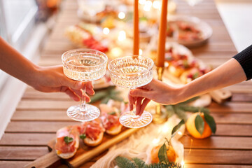 Hand with a glass of champagne in the foreground. A small table is served with snacks, bruschettas, and canapes. A decorated dining table with champagne glasses, candles and christmas tree an garland