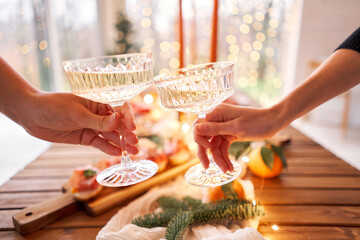Hand with a glass of champagne in the foreground. A small table is served with snacks, bruschettas, and canapes. A decorated dining table with champagne glasses, candles and christmas tree an garland