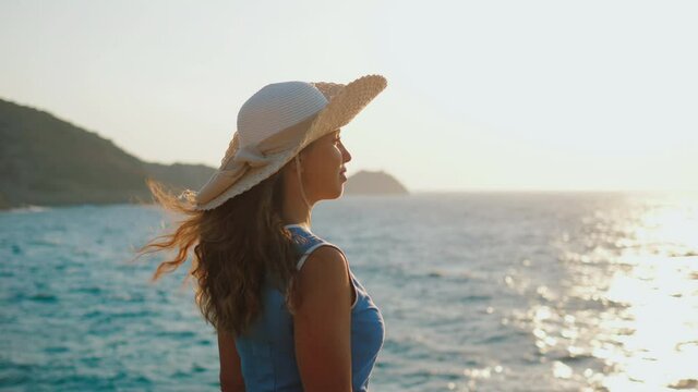 Attractive young woman in blue summer dress, straw hat standing on seashore washed by wave at beautiful sunset, admiring picturesque seascape, Looking into distance. Sea surf, slow motion