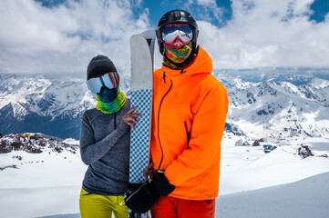 Young couple of skiers man and woman unrecognizable in snowboarding equipment masks on the background of snow-capped mountains high at a ski resort on a sunny day