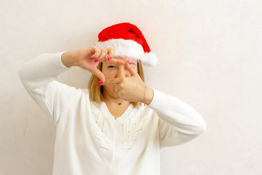 Body positive. Plus size girl with short hair stands against a light wall in a Santa Claus Christmas hat, fooling around and as if taking pictures on her fingers