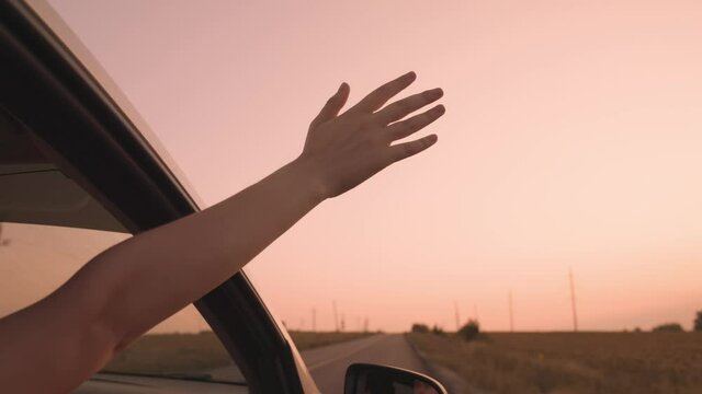 Free girl stretches out her hand from car in a pink sunset, catching wind with her hand while by transport on road, happy family, going on dream adventure vacation, having fun on road trip