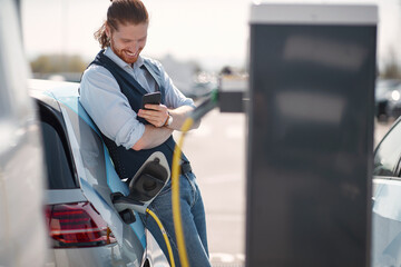 Smiling man using smartphone near electric car on charging station