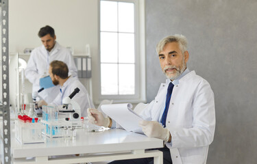 Portrait of a senior researcher working with various trace elements in a modern medical laboratory. Serious man in a white coat sits with notes on the clipboard and reads a sample analysis report.