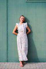 vertical portrait of a beautiful woman leaning on an industrial green wall. She is wearing a summer dress