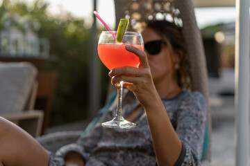 Young woman sitting on a rocking chair holding a colored glass with a fruit cocktail during a party...