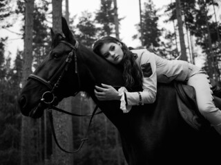 A young beautiful brunette rider in an elegant retro suit sitting astride a black horse leaning...