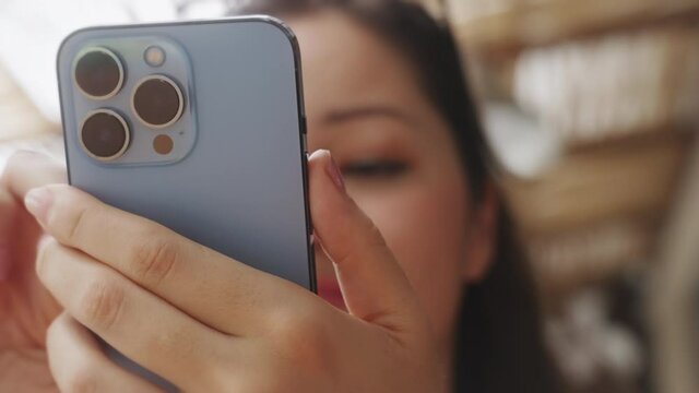 Dolly shot. A beautiful Asian blogger girl holds a new fashionable phone in her hands and sees a pleasant message and smiles while sitting in a sunny summer cafe. A close-up women. Slow motion.