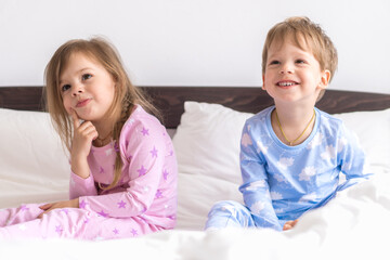 Two Preschool Toddler Children Siblings Boy And Girl Smiling Look At Side In Pink Blue Pajamas On White Bed. Little Baby Twins Have Fun. Happy Kids On Quarantine At Home. Friendship, Family Concept