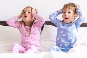 Two Preschool Toddler Children Siblings Boy And Girl portray horror surprise In Pink Blue Pajamas On White Bed. Little Baby Twins Have Fun. Happy Kids On Quarantine At Home. Friendship, Family Concept