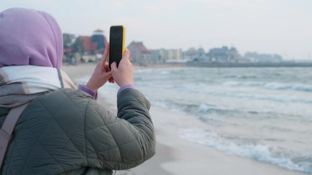 Tourist woman in warm clothes does photos by smartphone on the winter beach. Traveller at the cold sea coast in the off-season, 4k 60p