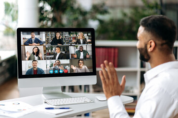 Video call, online conference. Over shoulder view of Indian man, on computer screen, talks with multinational group of successful business people, virtual business meeting, telecommunication concept