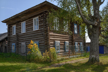 Fototapeta na wymiar An old two-storey residential log house with a front garden and flowers in a traditional Russian style in summer. Settlement Nyrob (Northern Ural, Russia). Green grass and a tall tree nearby 