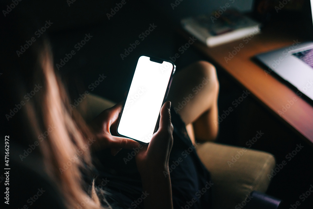 Wall mural woman holding a smartphone with a white screen mock up, resting on armchair in living room in the ni