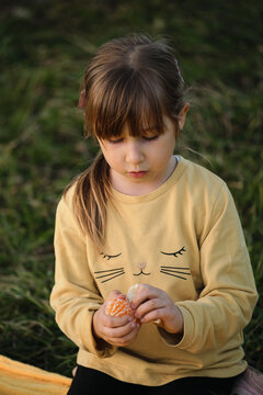 Child with brown hair and bangs is focused. Little Caucasian girl of five years old is sitting in park on blanket and cleaning tangerine with two hands.
