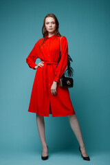 High fashion photo of a beautiful elegant young woman in a pretty red dress, black handbag posing over turquoise, blue background. Studio Shot.