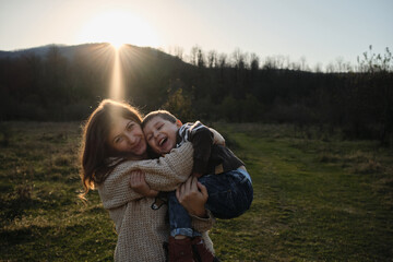 Spend childhood in nature with beloved mother. Joy of motherhood. Warm setting sun. Caucasian boy in striped sweater is sitting merrily in arms of woman and they are both laughing hugging.