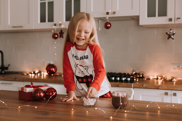 blonde little girl in red Christmas pajamas adds marshmallows to a mug with a hot drink in a beautifully decorated kitchen for the holiday. lifestyle. space for text. High quality photo