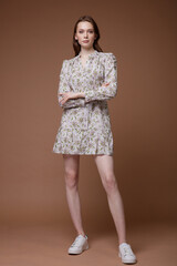 High fashion photo of a beautiful elegant young woman in a pretty short beige shirt dress with floral patterns, white sneakers posing over brown background. Studio Shot.