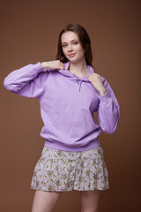 Obraz premium High fashion photo of a beautiful elegant young woman in a pretty short beige shirt dress with floral patterns, white sneakers, purple sweatshirt posing over brown background. Studio Shot. Portrait