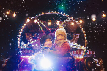 Fototapeta na wymiar Little cute kids - girl and boy attend the fun fair in the evening at the snow - Christmas market