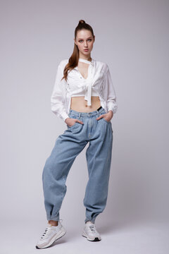 High fashion photo of a beautiful elegant young woman in a pretty white shirt and sneakers, blue denim jeans posing over white, soft gray background. Studio Shot.