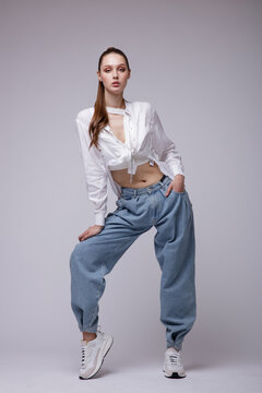 High fashion photo of a beautiful elegant young woman in a pretty white shirt and sneakers, blue denim jeans posing over white, soft gray background. Studio Shot.