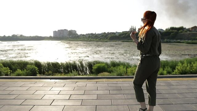 a beautiful red-haired woman in a khaki jumpsuit walks along the embankment in the park at sunset