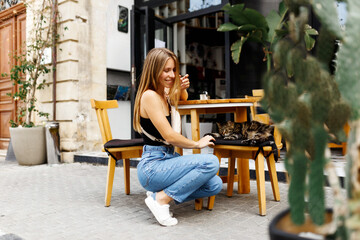 A young hipster woman sits at a table at an outdoor cafe bar and plays with the cat. Summertime .