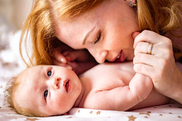 Happy redhead mother hugging little newborn baby lying together on cotton bed enjoying motherhood