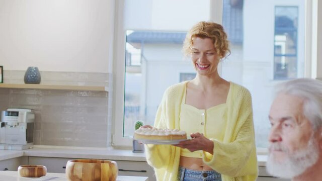 Blonde curly woman bringing tasty pie to the table for her family. Senior man smiling with pleasure. Happy family gathering together