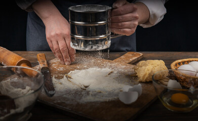 Women's hands, flour and dough. A woman in an apron cooking dough for homemade baking, a rustic home cozy atmosphere, a dark background with unusual lighting.