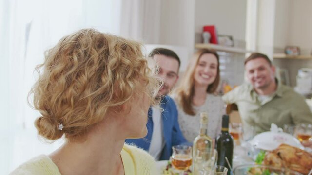 Medium shot of young caucasian woman holding camera in her hands and making video call with her diverse friends sitting at dining table, looking at camera and waving hands