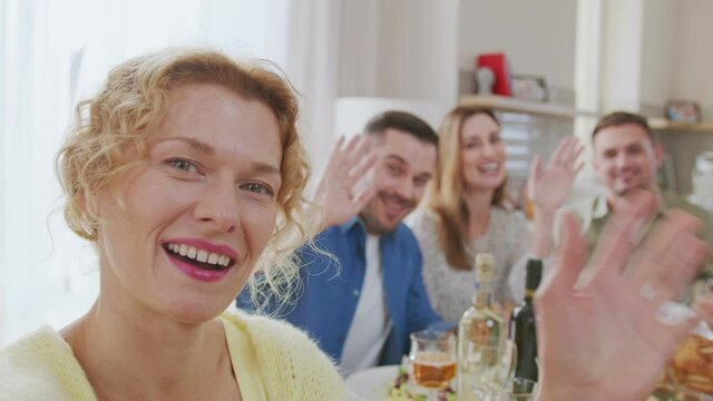 Medium shot of young caucasian woman holding camera in her hands and making video call with her diverse friends sitting at dining table, looking at camera and waving hands