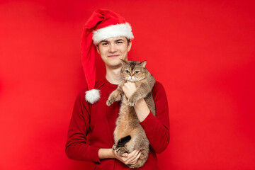 Young man in Santa's cap holds a cute British cat on a red background.