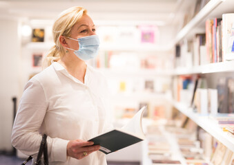 Handsome mature female in protective face mask choosing new books on shelves in museum shop