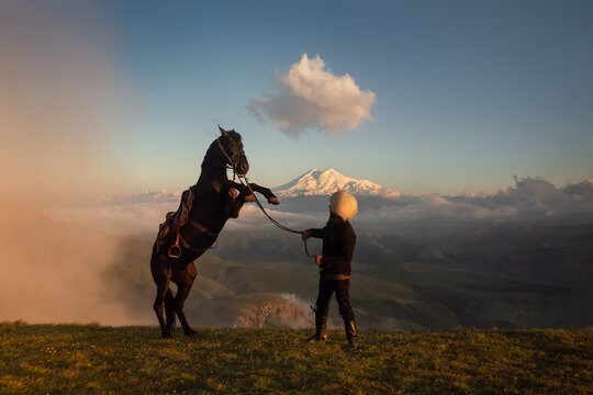 An epic view of the largest mountain in Europe, Elbrus. In the foreground is a Caucasian man in a fur hat and a rearing horse. Mountain horse of the Karachai breed. Bermamyt plateau. North Caucasus.