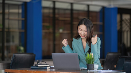 Happy excited successful Asian young businesswoman triumphing with laptop at modern office