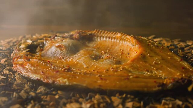 Delicatessen ide fish with spices smoked on wooden shavings and charcoals with rising up steam at bright light in smokehouse closeup slow motion