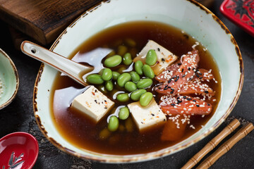 Middle close-up of miso soup with salmon fillet, edamame beans and chunks of tofu cheese served in...