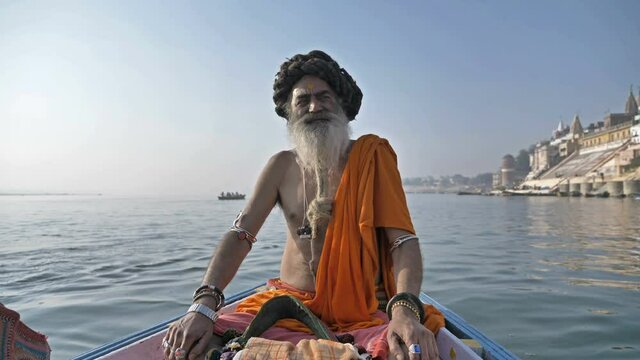 A bearded old or elderly male sadhu or Hindu priest wearing traditional clothes is sitting on a wooden boat in the Ganges River and looking at the camera while chanting a mantra or prayer in Varanasi