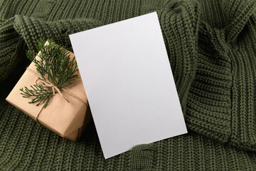 empty 5x7 card mockup kraft gift box with fir decoration on knitted green sweater, design element for wedding invitation, thank you or greeting card. Christmas winter card mock up background