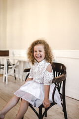 a beautiful little curly-haired girl sitting in the middle of a large room on a chair. smiling happily