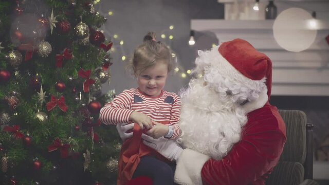 Adorable child sitting on Santa Claus lap near christmas tree. Real time