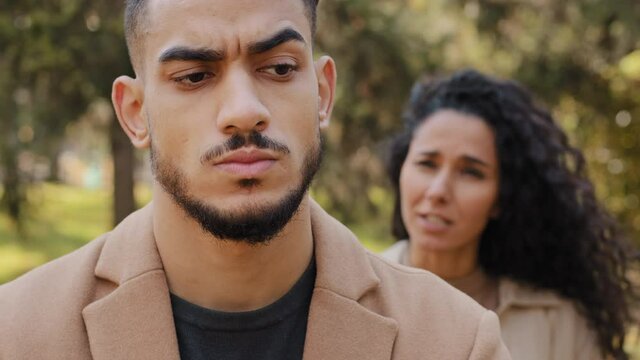 Emotional quarrel of hispanic couple outdoors angry girlfriend in background yelling and scolding boyfriend upset bearded man in foreground not listening reproaches unhappy marriage of young people.