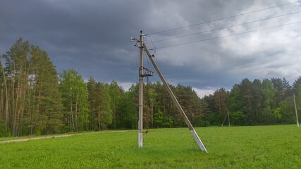 Fototapeta na wymiar A reinforced concrete pole of a power line with electric wires stands in a meadow near the forest. A thunderstorm is coming from behind the forest.