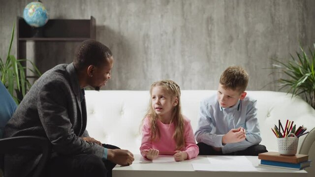Little girl pupil answers nodding African-American tutor while classmate sits on sofa at home English lesson with native speaker slow motion