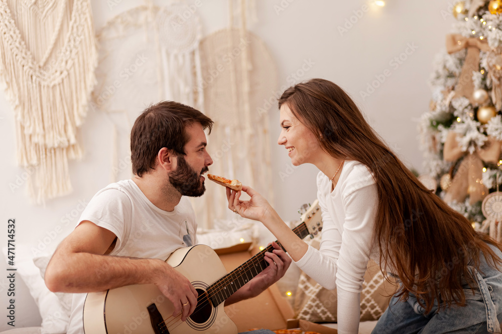 Wall mural Beautiful happy loving couple celebrating new year at home, having fun, eating pizza. Man playing the guitar, woman smiling. Romantic atmosphere, cozy interior, family time