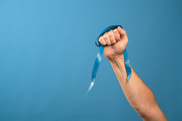 Sports and a healthy lifestyle, a figure without excess weight and good health. A man's hand holds a centimeter ribbon in his fist, blue background