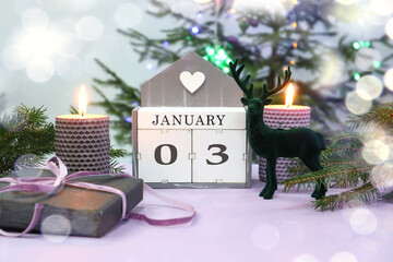 Calendar for January 3: the name of the month in English, numbers 0 and 3, burning candles, a gift tied with a purple ribbon, a figurine of a deer against the background of a festive tree, bokeh